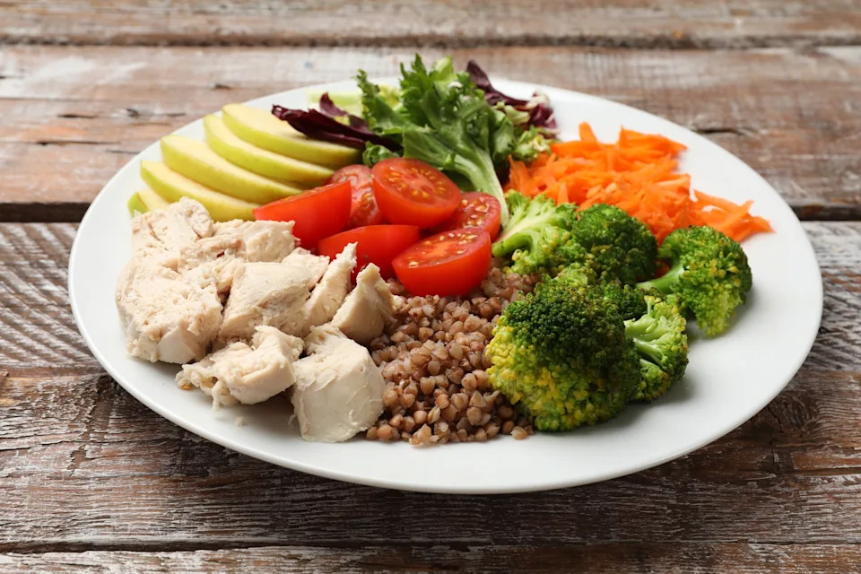 Plate with chicken, buckwheat, broccoli, tomatoes, apple slices, greens, and grated carrots on a wooden table