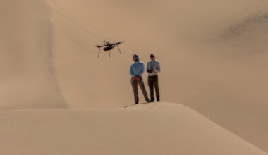 Two people stand side by side on a sandy hill, or dune. The person on the left is standing in a blue top, while the person on the right in a gray top is holding a controller. Above and to the left of their heads is a rotorcraft flying above the dune. The background of this image is more sandy dunes.