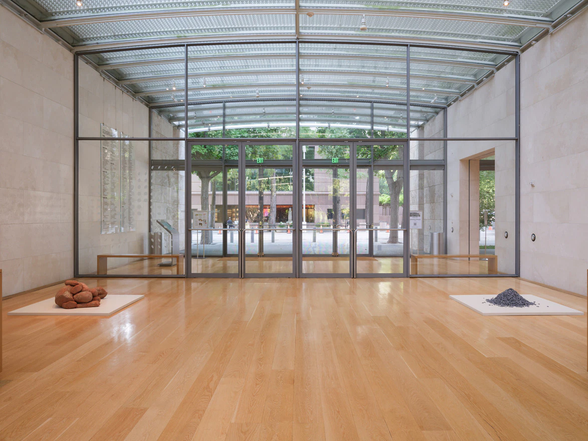 Modern art gallery showcasing minimalist installations on wooden flooring with large glass windows overlooking a garden.