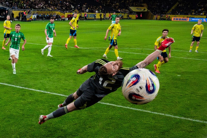 A goalkeeper dives to his left, fully outstretched, attempting to save a soccer ball as players in yellow and green jerseys watch intently during a match in a crowded stadium.