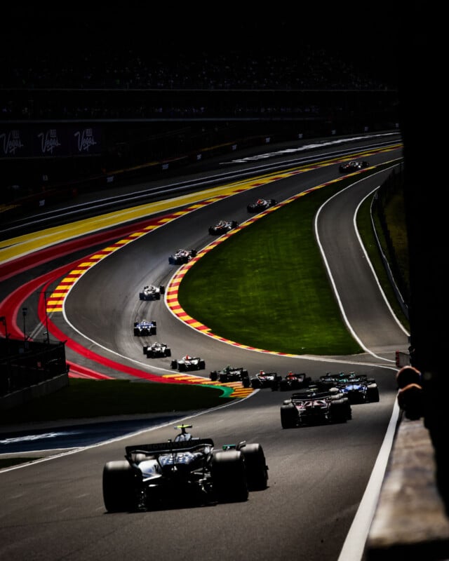 Formula 1 cars race closely together on a winding, brightly colored track with dramatic lighting and shadows, surrounded by green grass and distant spectators in the stands.