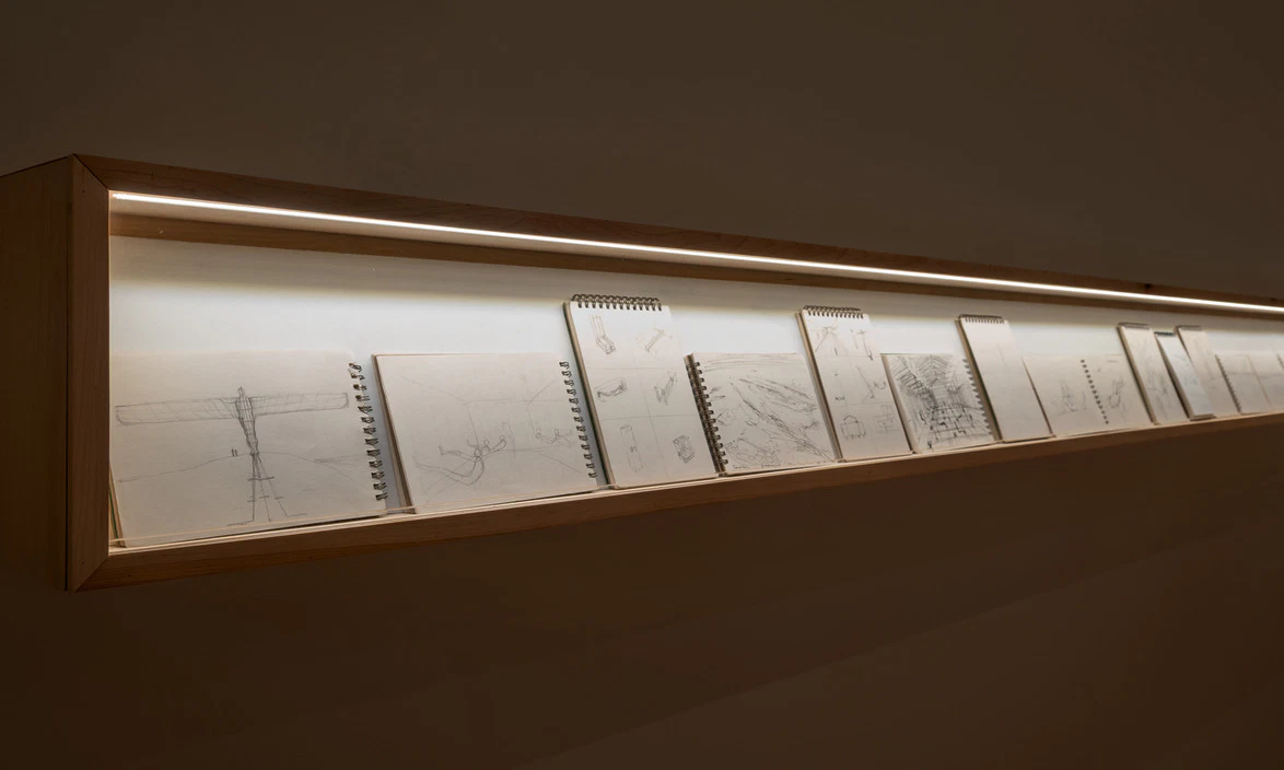 Art gallery display with several sketchbooks in a lit wooden case, showcasing various pencil drawings on paper.