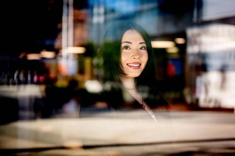 An Asian young woman smiles surrounded by blurry reflections in a window.