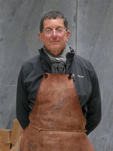 Man wearing apron and scarf stands in workshop with cardboard boxes in background