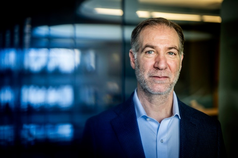 A man in a blue shirt and dark blazer stands in front of a blurry wall of windows.