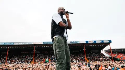 Stewart Baxter A side shot of Chiedu Oraka on stage. He is bald and is wearing black-rimmed glasses. He is also wearing a white short-sleeved t-shirt, a black bodywarmer and camoflauged trousers. He is holding a black microphone with his left hand to his mouth. Crowds of people can be seen in the stadium in the background. The sky is white. 