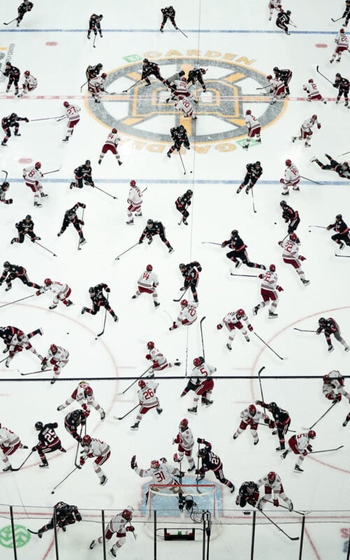 Overhead view of two ice hockey teams wearing dark and white uniforms practicing on the rink, with many players skating and handling pucks before a game. The arena logo is visible at center ice.