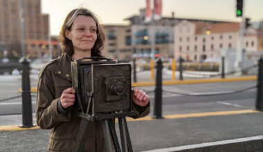 Game designer Susannah Emery stands behind a statue of a camera. She is a white woman with brown eyes and brunette hair. She is wearing glasses and a muted green jacket.