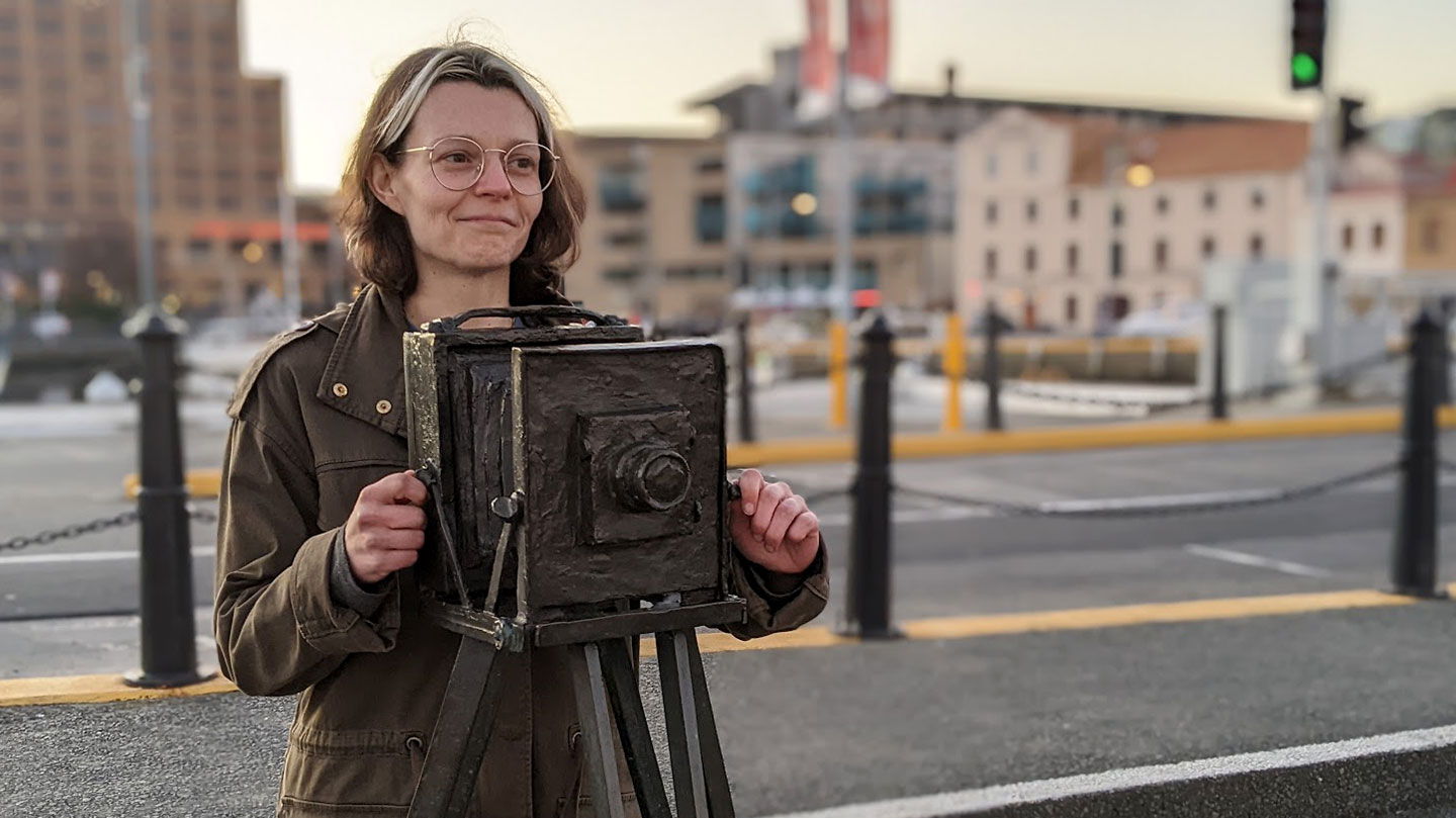 Game designer Susannah Emery stands behind a statue of a camera. She is a white woman with brown eyes and brunette hair. She is wearing glasses and a muted green jacket.
