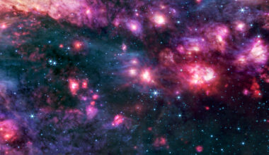 Cosmic clouds of pink and purple, some with bright centers, are surrounded by dark areas that appear like black space dotted with bright blue stars. A group of small clouds to the right is more red than any other area of the image.
