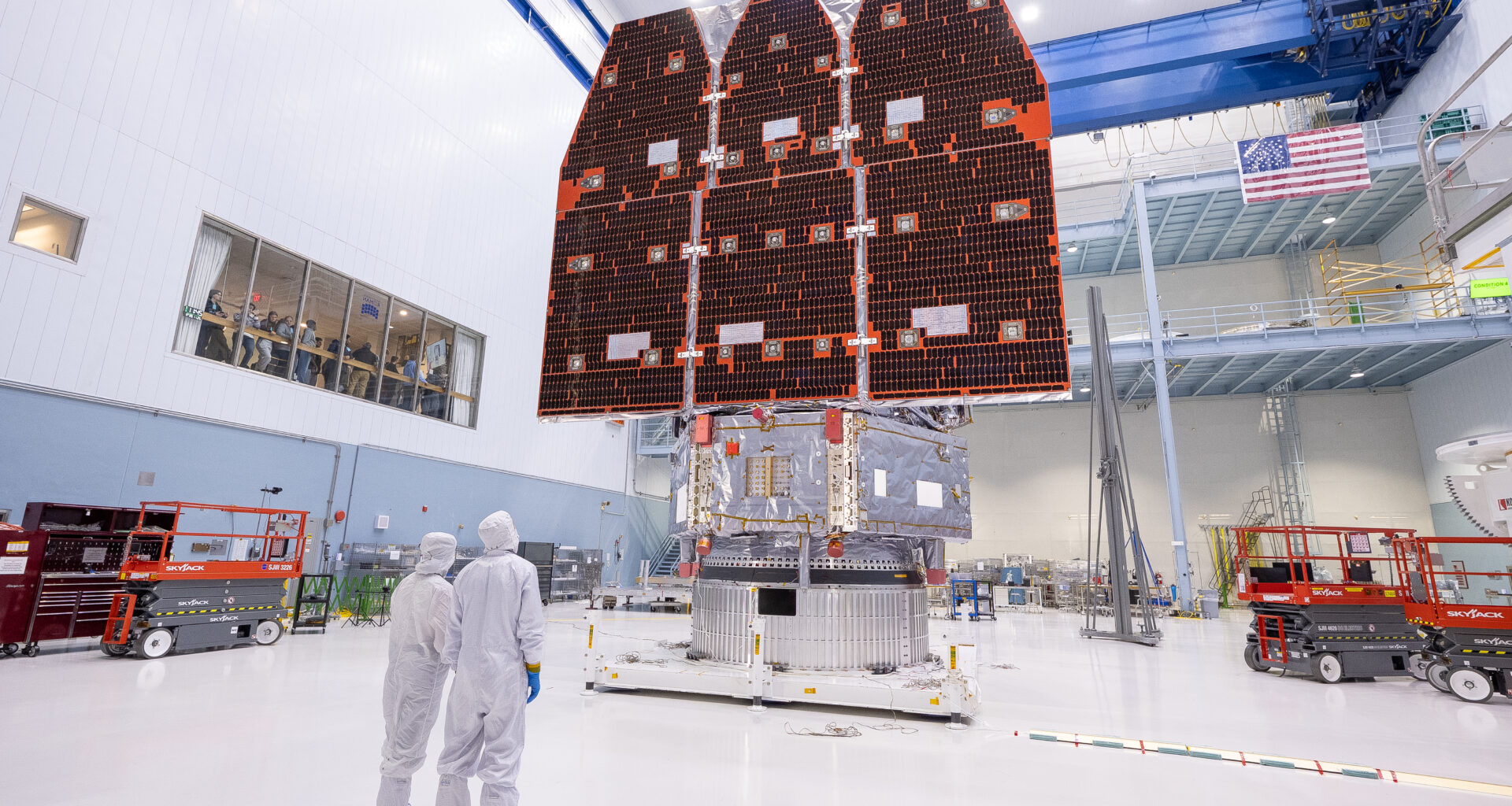Two people in white jumpsuits look up at a large telescope with three black and orange panels facing them. They are in a large white cleanroom with equipment and scaffolding.