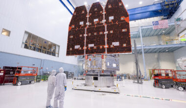 Two people in white jumpsuits look up at a large telescope with three black and orange panels facing them. They are in a large white cleanroom with equipment and scaffolding.