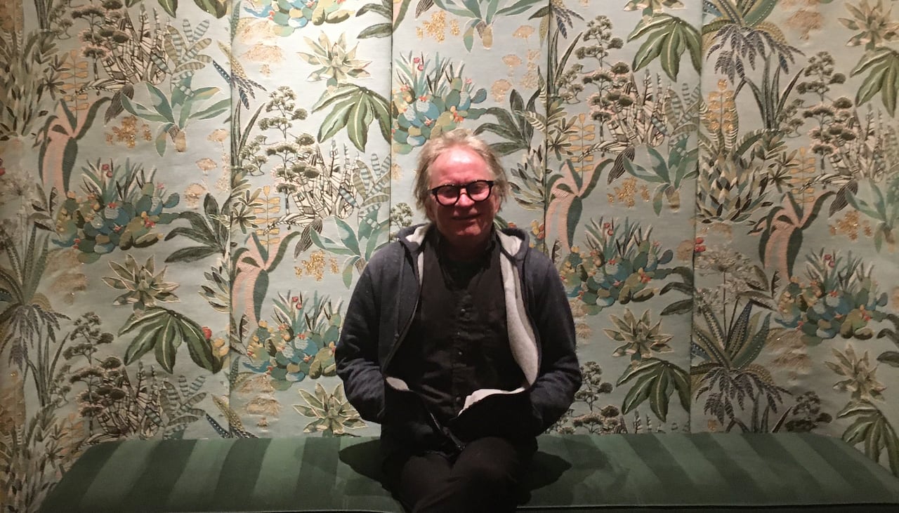 A man sits on a green bench, against colourful floral wallpaper, smiling at the camera