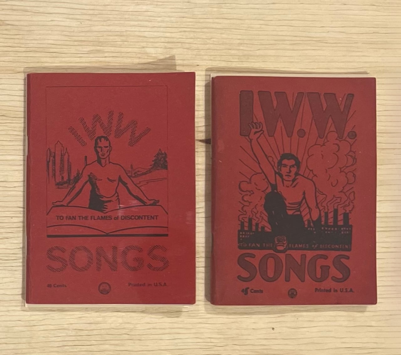 Two small red books sit on a wood surface. Both covers show a male figure and bear the words "IWW Songs" as well as the line "To fan the flames of discontent."