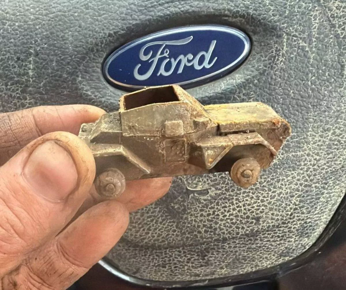 An individual is holding a miniature brass model of a vintage Ford vehicle, positioned against a backdrop featuring the Ford logo.