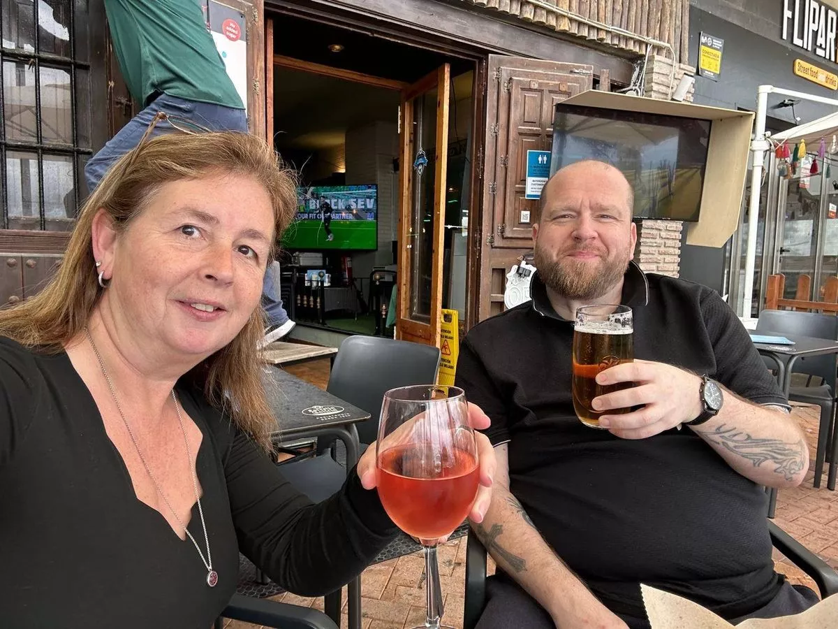 Two individuals, a man and a woman, are seated at a table outside a restaurant. They are both holding glasses of a red beverage and appear to be engaged in a social setting. The background features the entrance of the establishment and a partially visible outdoor seating area.