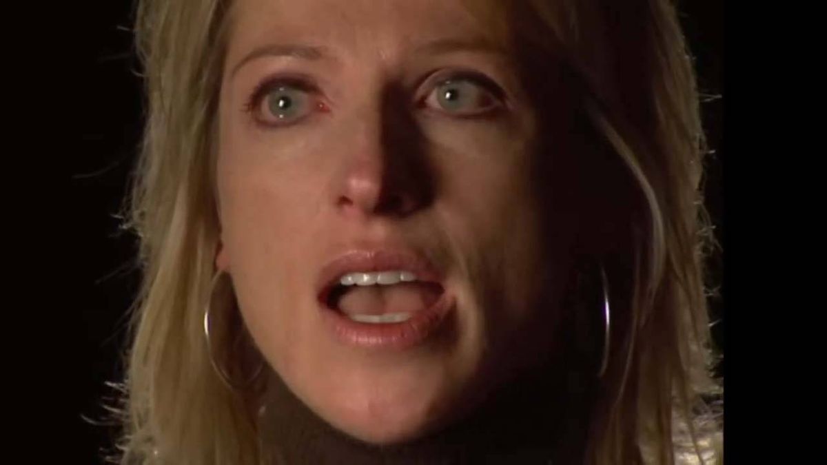 A close-up portrait of an individual with long, light-colored hair, appearing to speak or express surprise, with the face partially illuminated against a dark background.