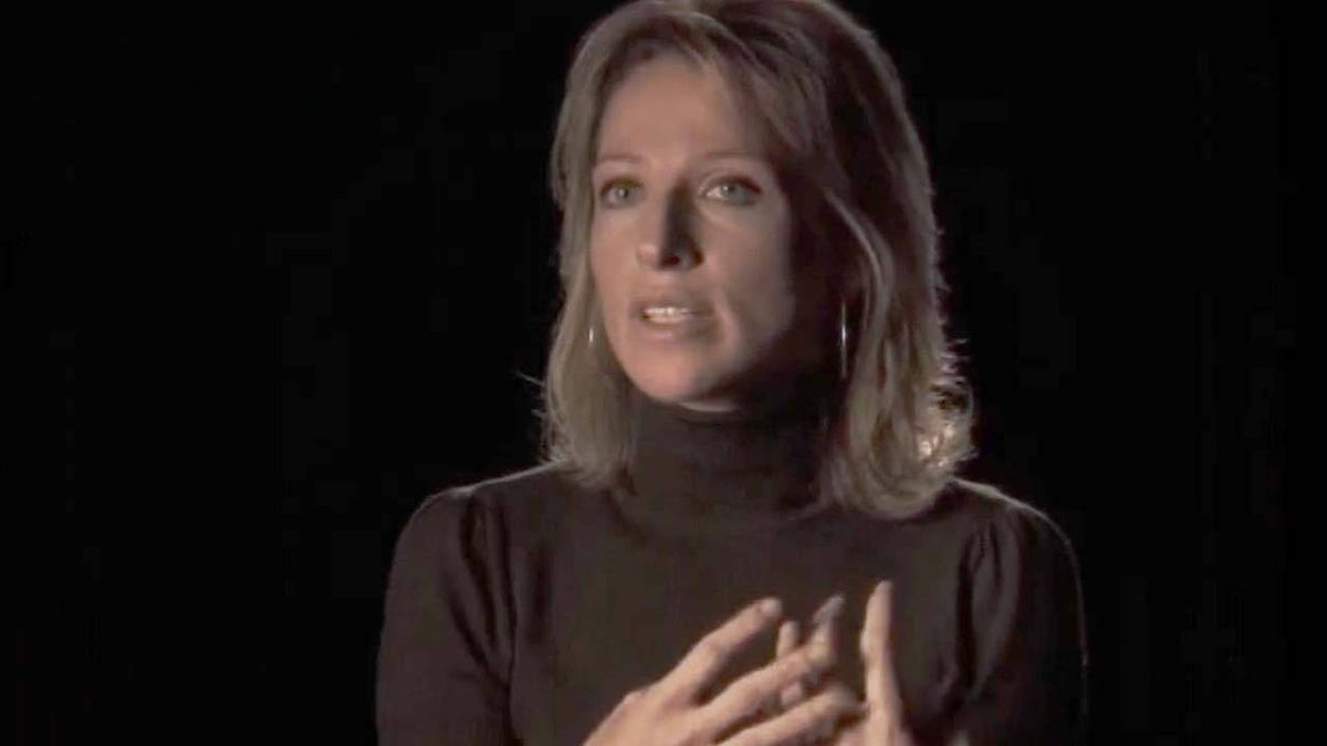 An individual with long, light-colored hair, dressed in a dark, high-necked garment, appears to be speaking earnestly while placing hands on their chest in a focused manner against a darkened backdrop.