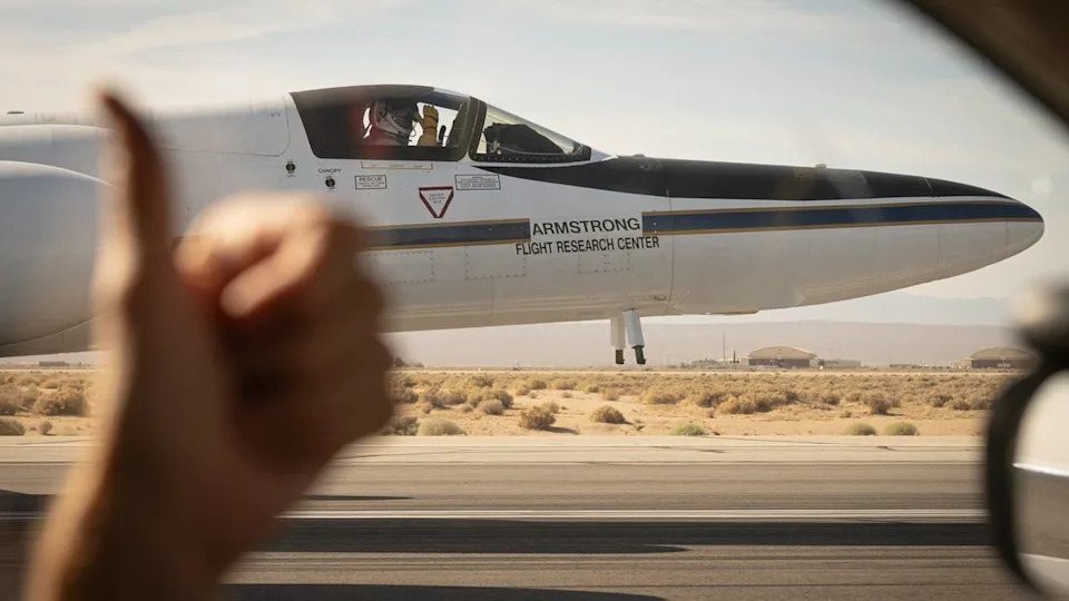 A view from the driver's side window of a car shows a white plane on the runway where the driver's hand shows a thumbs up on the left side of the image.