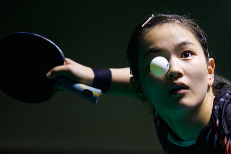 A table tennis player intensely focuses as a ping pong ball appears to hover directly in front of her eye. She holds a paddle, ready to hit, with a look of concentration on her face.