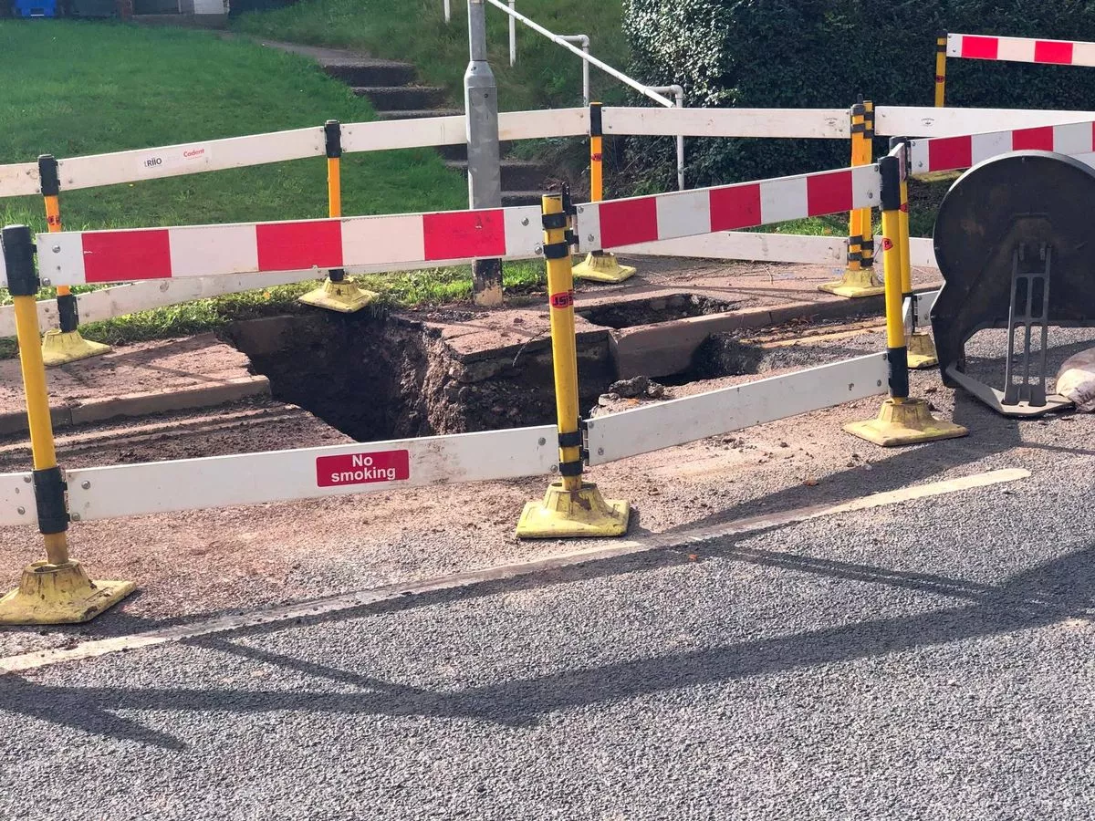 A section of a road cordoned off with yellow and white bollards, featuring red and white stripes. The area indicates a site of ongoing construction or maintenance, with visible roadwork evident. The surrounding environment includes a grassy field and distant structures.
