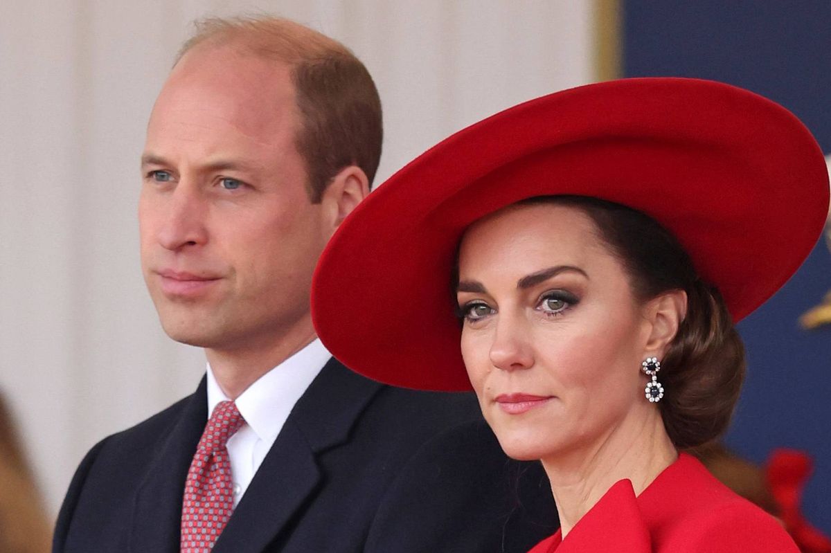 An individual in formal attire, likely a public figure, stands next to a woman wearing a red hat and a red dress, both individuals appear to be engaged in an official event or ceremony.