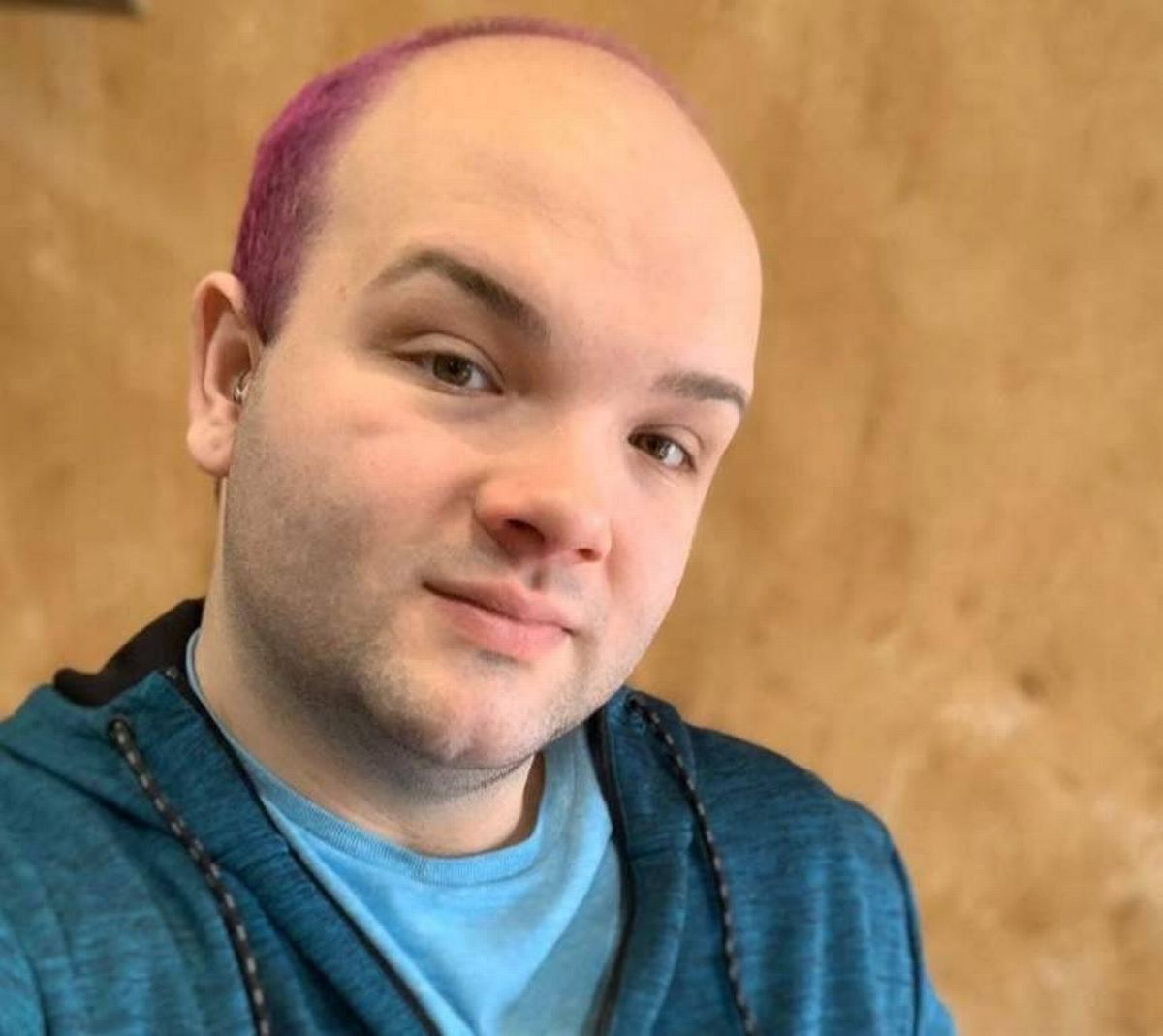 An individual dressed in a blue hoodie and a light blue shirt is captured in a close-up portrait, standing against a textured wooden background, and appearing to be engaged in a thoughtful expression.