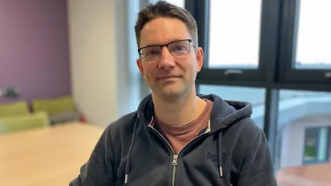 BBC/Seb Cheer A man with short brown hair and glasses sits in a meeting room and smiles at the camera.