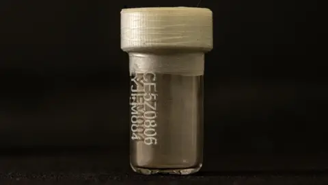 Tony Jolliffe/BBC News A small clear vial with a white plastic top and tape around the lid. It is lit against a black background. On the front is a code. Inside at the bottom there is a coating of dust.