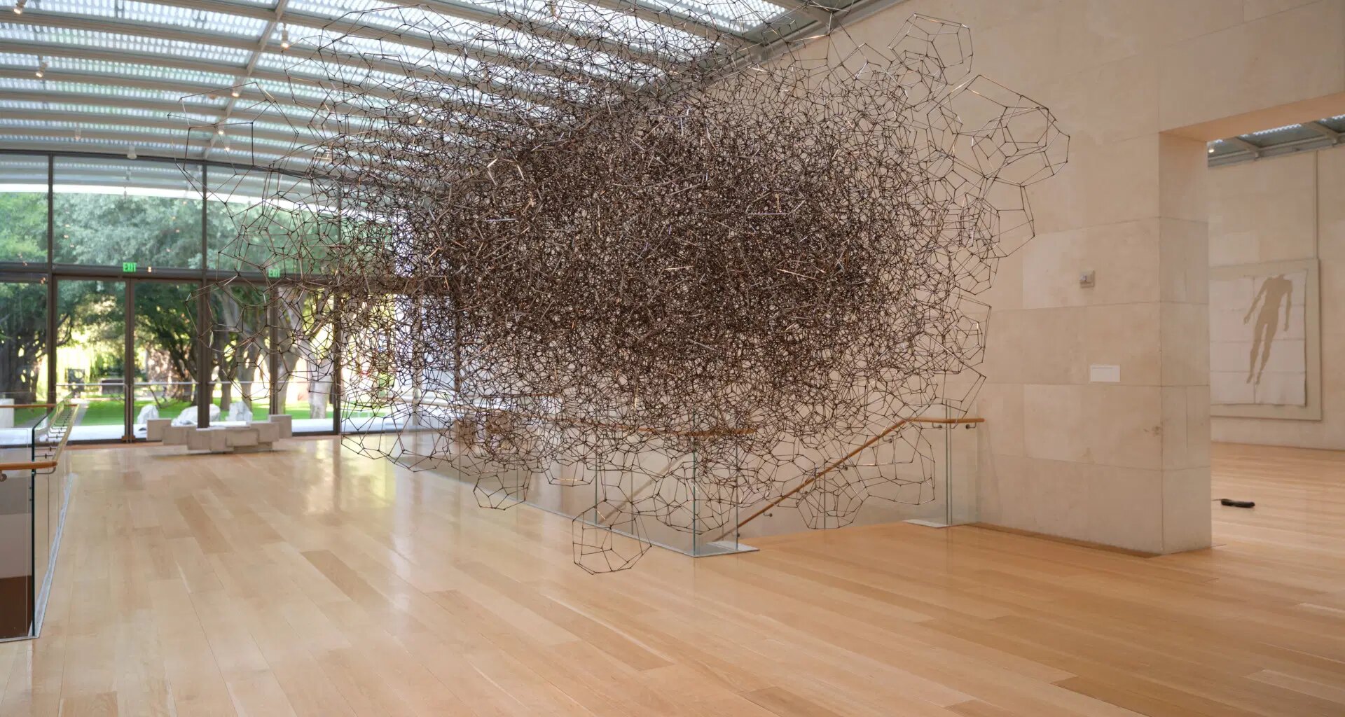 Large intricate metallic sculpture suspended in a spacious, sunlit gallery with wooden floors and glass walls.