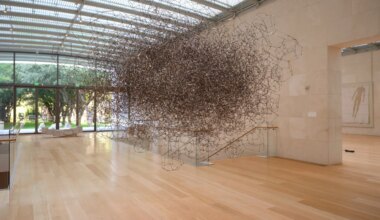 Large intricate metallic sculpture suspended in a spacious, sunlit gallery with wooden floors and glass walls.