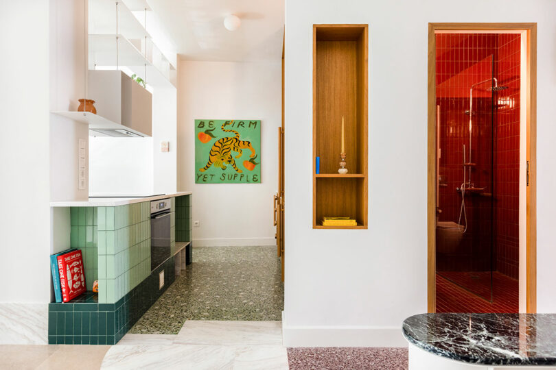 Modern kitchen with green tiles, open shelving, and a visible red-tiled shower room. A colorful "Be Firm Yet Supple" tiger artwork hangs on the far wall, giving this space a vibrant touch 