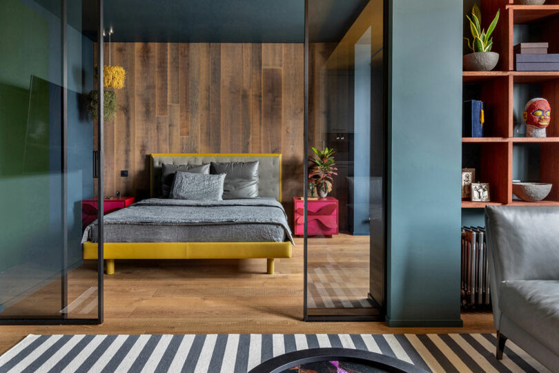 A modern bedroom with a wooden accent wall, yellow bed frame, gray bedding, glass doors, and striped rug