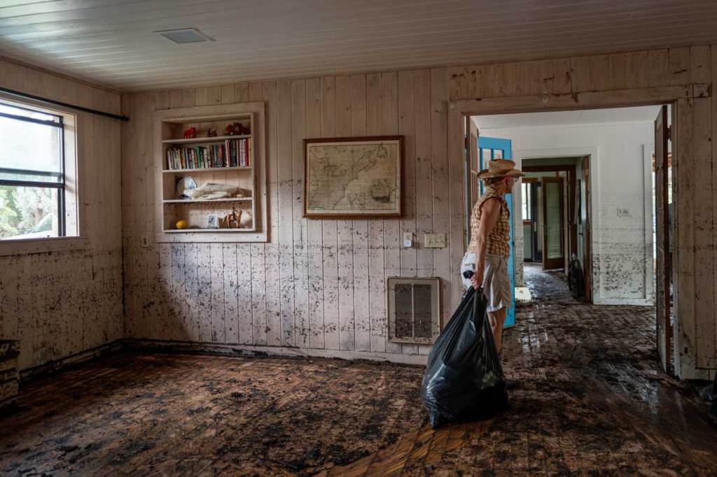 A photo of a woman dragging a full garbage bag through a home that has been flooded. It has no furniture, and mud is smeared on the floor and walls.
