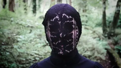 Risteárd Ó hAodha  Person standing on a forest path surrounded by dense greenery, wearing a dark outfit and lace-like head covering; face intentionally blurred for anonymity.