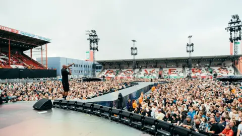 Stewart Baxter A wide-angle photo of Chiedu Oraka on stage facing away from the camera but towards the crowds of people that fill the stadium. He has his left arm pointing in the air and is holding a black microphone to his mouth with his right hand. He is wearing a black t-shirt and black shorts. The sky is white. 