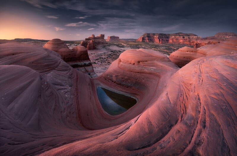 A dramatic desert landscape at sunset features swirling red sandstone formations and a small, heart-shaped pool of water nestled between the rocks, with distant mesas and a glowing sky in the background.