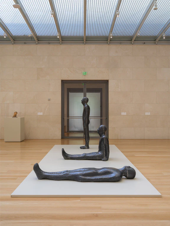 Three dark statues in various poses displayed on a white platform in a modern art gallery setting.