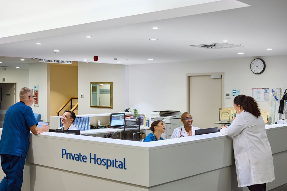 Medical staff interacting at hospital reception. Surgeon in blue scrubs using tablet while doctors and nurses smiling and talking. Bright and modern reception area with computers, paperwork, and equipment.