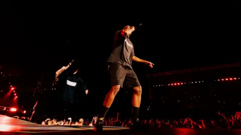 Anna Lee Media A photo of Chiedu Oraka on stage, taken from the crowd. He is holding a black microphone to his mouth with his right hand. He is wearing a black t-shirt with white branding, black knee-length shorts, black socks and black trainers. Red lights can be seen on stage and across the stadium. People can be seen in the crowd behind him. The night is dark and the sky is black. 