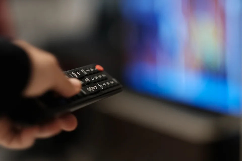 Person holding a TV remote, pointing it at a flat-screen TV in a living room setting