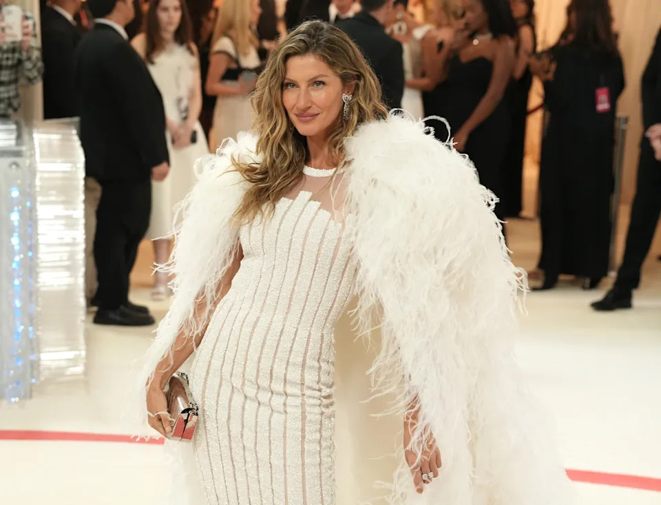 A person on a red carpet wearing a white dress with feathered cape, holding a clutch, surrounded by event attendees