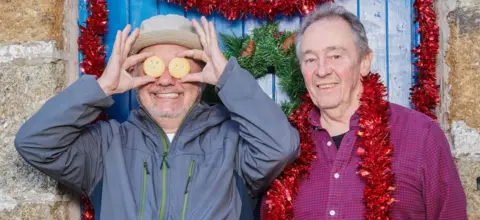 Bob Mortimer and Paul Whitehouse standing in front of a house with tinsel around their necks