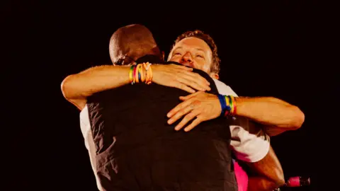 Anna Lee Media Chiedu Oraka facing away from the camera, with Chris Martin, who is facing the camera, embracing him. Oraka is wearing a white short-sleeved t-shirt and a black bodywarmer. Martin has colourful wristbands on both wrists and is smiling over Oraka's shoulder. The sky is black. 