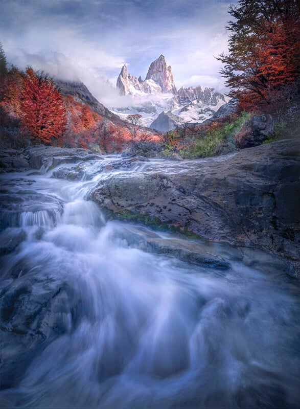 A rushing mountain stream flows over rocks, surrounded by vibrant red autumn trees, with snow-capped jagged peaks rising dramatically in the background under a partly cloudy sky.