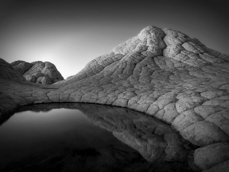 Black and white photo of a rocky, textured mountain with a small, still pool of water in the foreground reflecting the mountain’s surface. The landscape appears otherworldly and barren under a clear sky.
