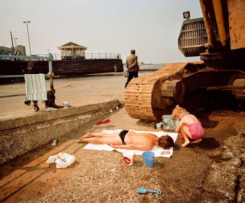 A person sunbathes on a towel near a large excavator, while a child in pink plays beside the machine. Sand toys are scattered on the ground, and a man walks away toward the seaside promenade in the background.