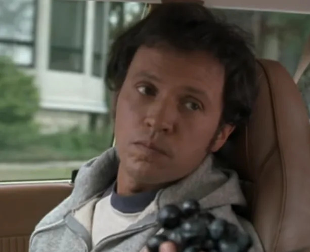 Person in a car seat looking sideways with a neutral expression, holding a cluster of grapes