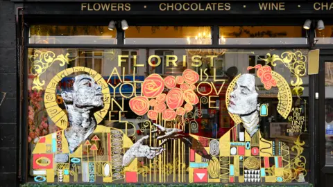 Ash Field/Our Tiny Windows A lavish window display in gold and red coloured paint takes up the entirety of a shop window. It shows two figures who are depicted as religious-looking icons reaching out their hands to each other with red flowers between them.