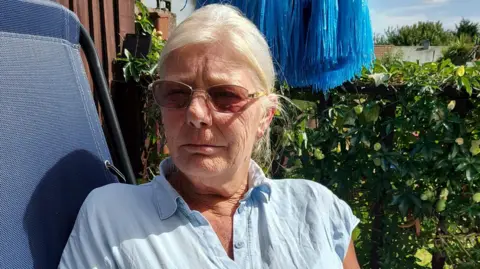 Wales News Service Belinda sitting in a outdoor blue char with a hedge behind her. She has light parted hair pushed back behind her ear and diamond ear rings. and is wearing a light blue shirt. 